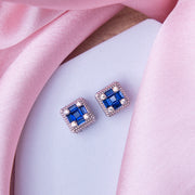 A pair of silver stud earrings with ultramarine blue gemstones, displayed on a pink fabric background.